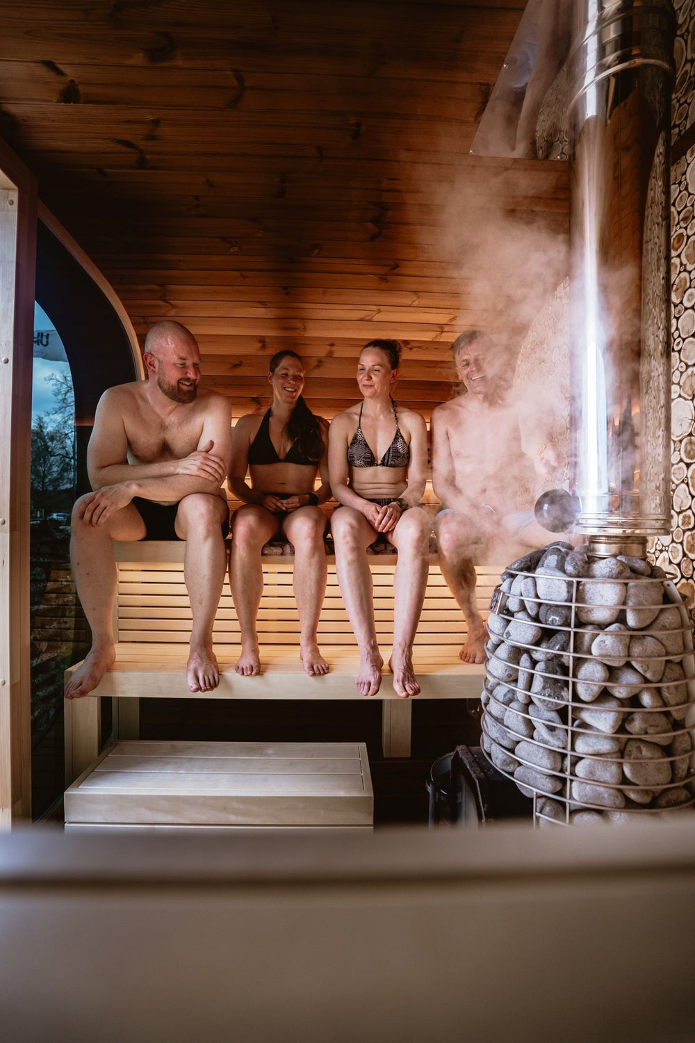 Four adults seated inside a timber sauna beside a stone-filled heater.