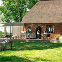 Wide garden view of the Burnout BBQ outdoor kitchen with guests dining on the terrace of a thatched cottage